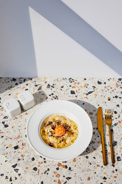 νόστιμα ζυμαρικά καρμπονάρα σερβίρεται με χρυσά μαχαιροπίρουνα, αλάτι και πιπέρι σέικερ σε πέτρινο τραπέζι στο φως του ήλιου - Φωτογραφία, εικόνα