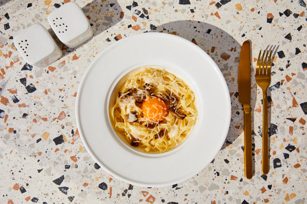bovenaanzicht van heerlijke pasta carbonara geserveerd met gouden bestek, zout en peper shakers op stenen tafel in zonlicht - Foto, afbeelding