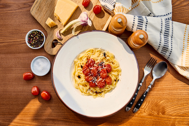 top view of delicious pasta with tomatoes served on wooden table with cutlery, napkin, seasoning and ingredients in sunlight - Foto, afbeelding