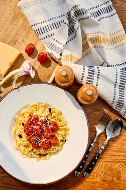 top view of delicious pasta with tomatoes served on wooden table with cutlery, napkin, seasoning and ingredients in sunlight - Photo, Image
