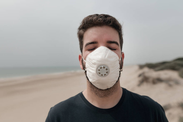 Jongeman op het strand draagt een medisch masker om ziektes en virussen te voorkomen. Coronavirus, epidemieën en allergie concept. Gezondheidsalarm status. Isolatiewoning. - Foto, afbeelding