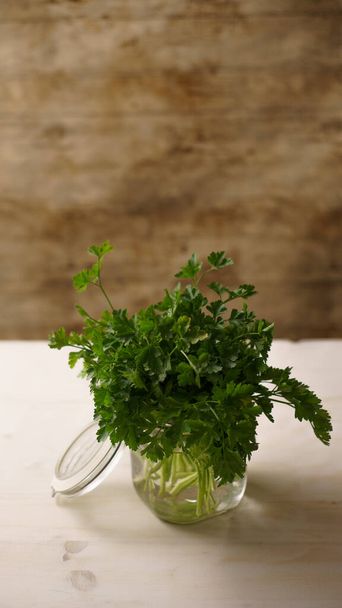 Vertical close up of an open glass jar full of water with a spring of green parsley in kitchen on light wooden background, mediterranean cuisine - Photo, Image