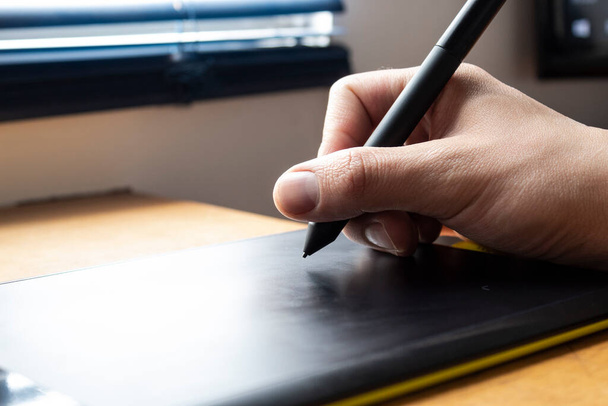 Close-up van een mannelijke hand die een zwart digitaal potlood over een zwart-groene tekentablet in bureau en thuiskantoor achtergrond houdt. Ideaal voor grafisch ontwerp, web en elke relatie met technologie - Foto, afbeelding