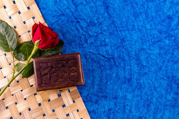 Draufsicht einer roten Rose über gewebten Bambusstreifen mit modelliertem braunem Kasten auf blauem Hintergrund. Klassisches Geschenkkonzept - Foto, Bild