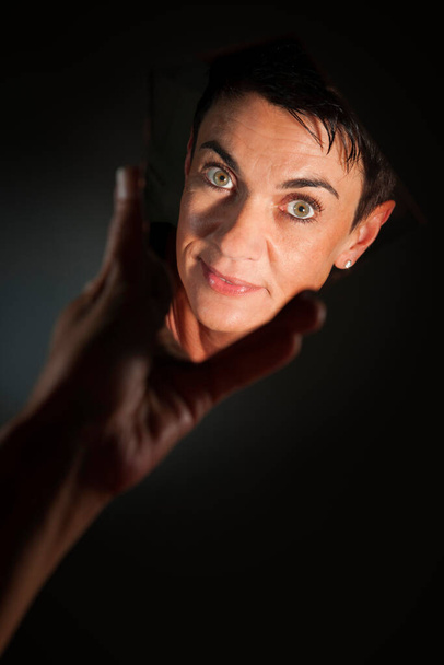 Gezichtsportret van een vrouw van middelbare leeftijd met zwart haar die in de camera kijkt in een hand gehouden spiegelscherf tegen een donkere achtergrond. - Foto, afbeelding