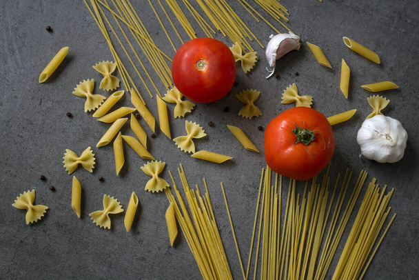 Still life with tomatoes and dry pasta of different types. Fresh ingredients top view. Dark grey background. Traditional cooking.  - Photo, Image