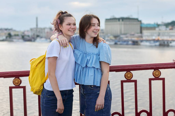 Portrait of two teenage girls standing on bridge over river, friends walk on summer sunny day. Friendship, lifestyle, youth, teens - Фото, изображение