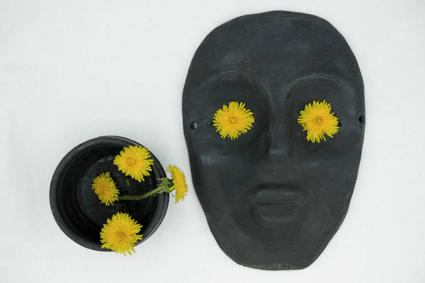 Still life with black ceramic mask and black cup on a white background. Yellow dandelion flowers in the eyes of the mask and in the cup. Mass produced mask. - Photo, Image
