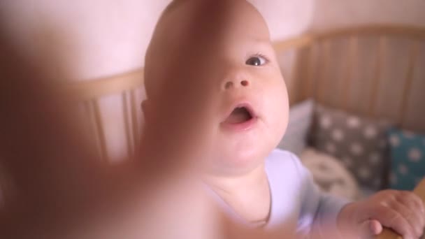 ベッドの中で面白い赤ちゃんが立ってカメラを見ています。子供は手をカメラに伸ばします。子供部屋の子供はベビーベッドにいる。子供はカメラに手を伸ばしてそれを調べる. - 映像、動画
