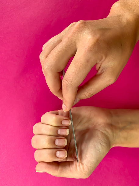 Mão de mulher da menina arquivando unhas com arquivo de unhas de metal em um fundo rosa. Auto-manicure em casa
 - Foto, Imagem