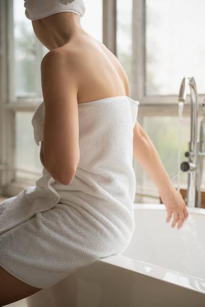 Woman wrapped in white towel sitting on the edge of bathtub and touching flowing water - 写真・画像
