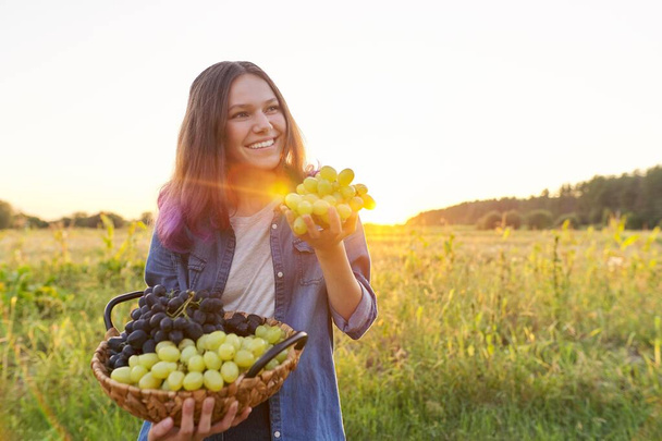 Beautiful smiling teenager girl with basket with crop of green and blue grapes, female on background of natural country landscape, sunset, copy space - Foto, immagini