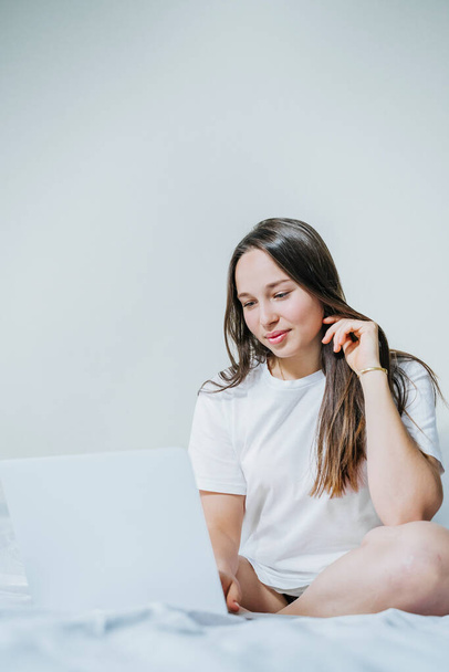 Young cheerful woman sitting on the bed using laptop for working from home. Space for text. - Foto, Bild