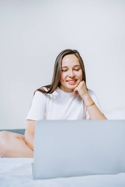Young cheerful woman sitting on the bed using laptop for working from home. Space for text. - Valokuva, kuva