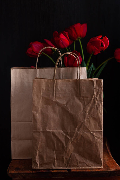 Kraft paper bags with blooming red tulips bouquet on black background. Dark moody low key minimalism style flowers and food delivery packaging banner copy space mockup. Donations help recycling boxes. - Photo, Image