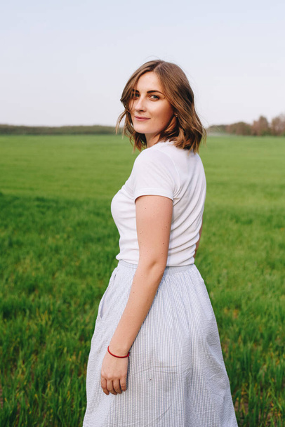 The girl is blonde, brown hair, in a white shirt and blue midi skirt. Walking in the field, through the green grass. Portrait of a girl. Positive and smile. - Foto, Bild