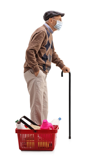 Full length profile shot of an elderly man with a protective face mask and a shopping basket full of groceries isolated on white background - Fotoğraf, Görsel