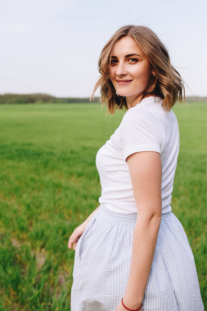 The girl is blonde, brown hair, in a white shirt and blue midi skirt. Walking in the field, through the green grass. Portrait of a girl. Positive and smile. - Фото, зображення
