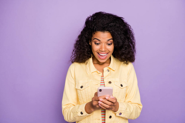 Portrait of crazy amazed afro american girl using her smartphone get social media notification typing blogging comment wear stylish yellow jacket isolated over purple color background - Foto, imagen