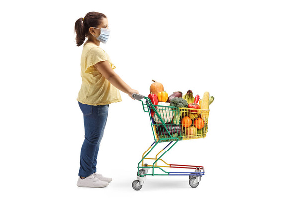 Full length profile shot of a girl standing with a shopping cart and wearing a medical face mask isolated on white background - 写真・画像
