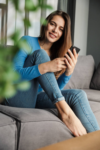 Bild der schönen jungen fröhlichen Frau mit Handy und lächelt, während sie auf dem Sofa im Wohnzimmer sitzt - Foto, Bild