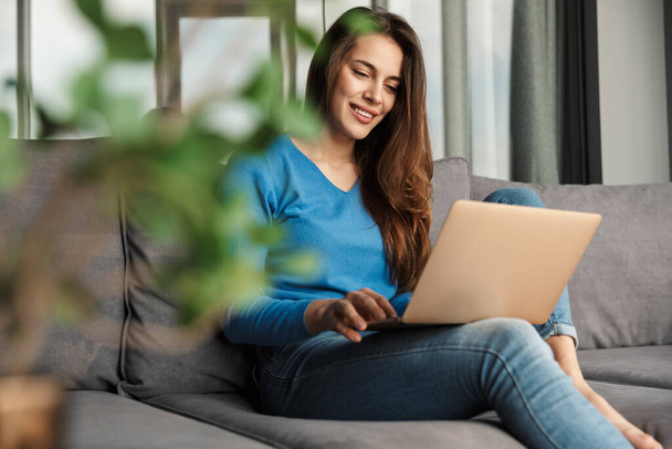 Image of seductive young happy woman using laptop and smiling while sitting on sofa at living room - Photo, image