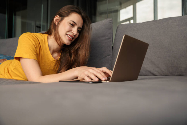 Image of smiling young cute woman using laptop and mobile phone while lying on couch at living room - Foto, Imagem