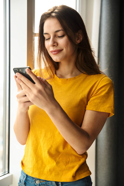 Image of pleased young cute woman using mobile phone and smiling while standing near window - Foto, imagen