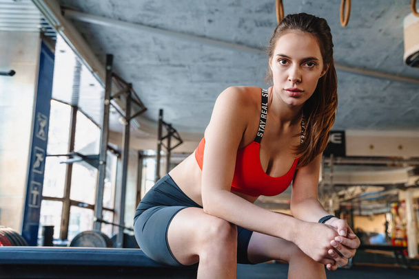Portrét mladé fitness sportovkyně dívající se do kamery a sedící při cvičení v atletické hale - Fotografie, Obrázek