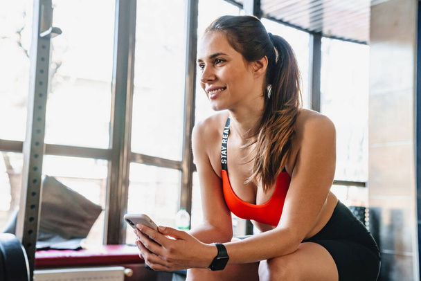 Portrait of joyful young sportswoman sitting and using cellphone while working out in athletic hall - Photo, Image