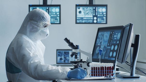 Scientist in protection suit and masks working in research lab using laboratory equipment: microscopes, test tubes. Coronavirus 2019-ncov hazard, pharmaceutical discovery, bacteriology and virology - Photo, image