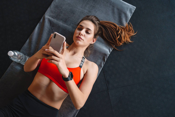 Vista dall'alto di una giovane sportiva attraente che utilizza il telefono cellulare mentre si trova su un tappeto fitness in palestra
 - Foto, immagini