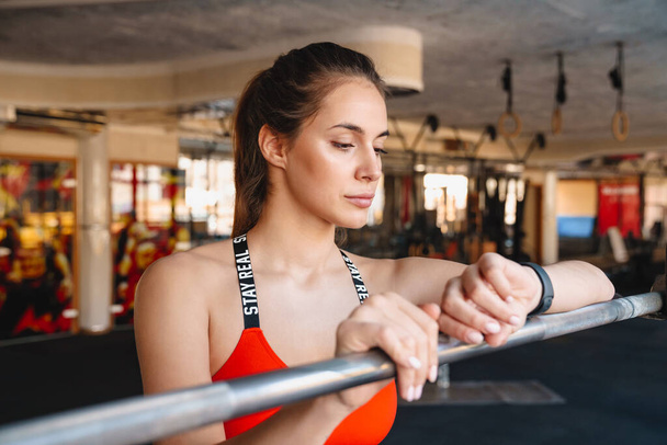 Attractive young fit sportswoman standing in the gym, resting after exercises, leaning on a rail, checking fitness watch - Φωτογραφία, εικόνα