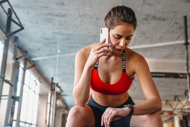 Portrait of focused young sportswoman talking on cellphone and using smartwatch while working out in athletic hall - Photo, image