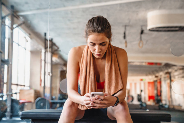 Attractive young fit sportswoman resting after exercises while sitting in the gym, using mobile phone - Foto, Imagem