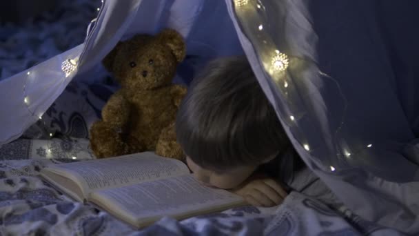 Little boy reads with pocket light. Toddler plays in tent made of linen sheet on bed. Cozy evening with favorite book. - Séquence, vidéo