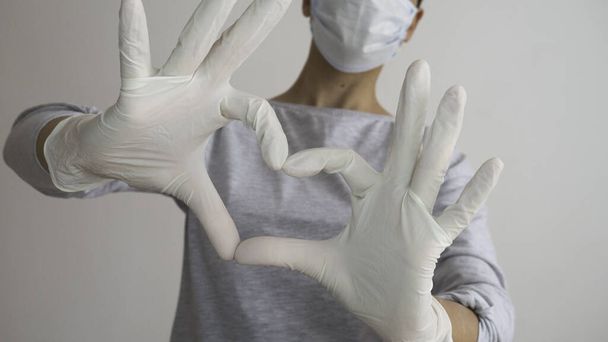 Γυναίκα με μάσκα προσώπου και τα χέρια σε latex λευκά γάντια δείχνει το σύμβολο της καρδιάς. - Φωτογραφία, εικόνα