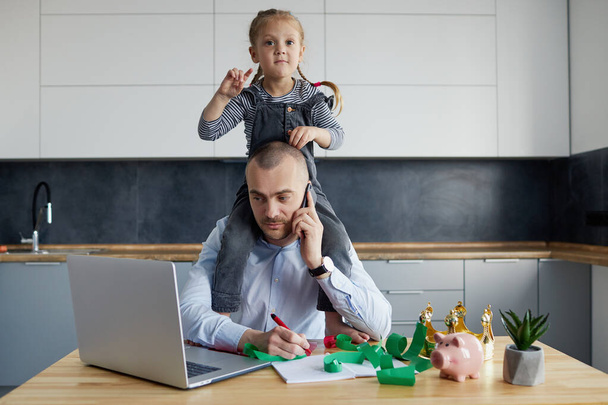 Father Working from home on laptop during quarantine. Little child girl make noise and distracts father from work on the kitchen office - Φωτογραφία, εικόνα