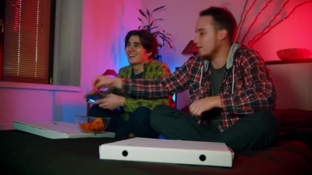 Dva mladí muži přátelé jíst chipsy a hrát hru s gamepady - jeden z nich prohrává - Záběry, video