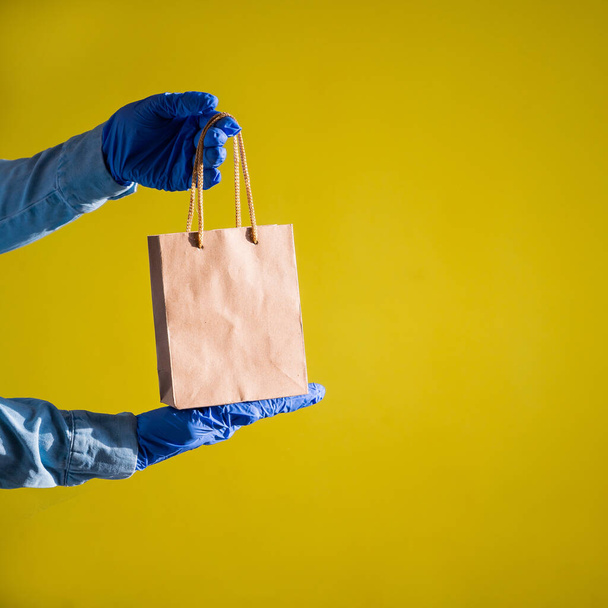 Closeup of female hands in gloves and a denim shirt. A delivery man is passes a small paper bag by the pens on a yellow background. Craft packaging for takeaway snack. Antimicrobial protection. - Foto, imagen
