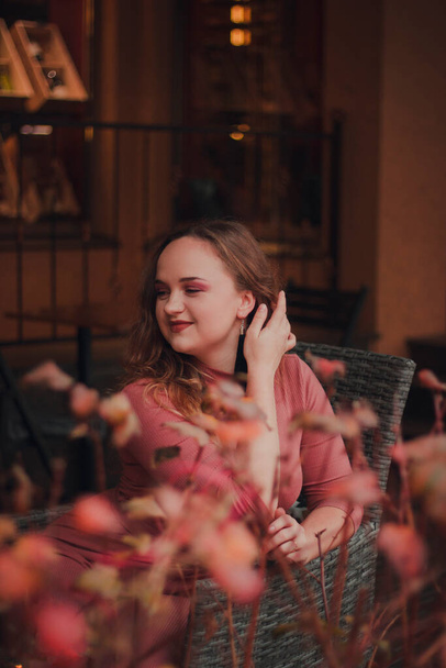 Een mooi, elegant, bescheiden meisje in een café op het terras. Gezellige warme sfeer. Lichte glimlach en bokeh op de achtergrond  - Foto, afbeelding