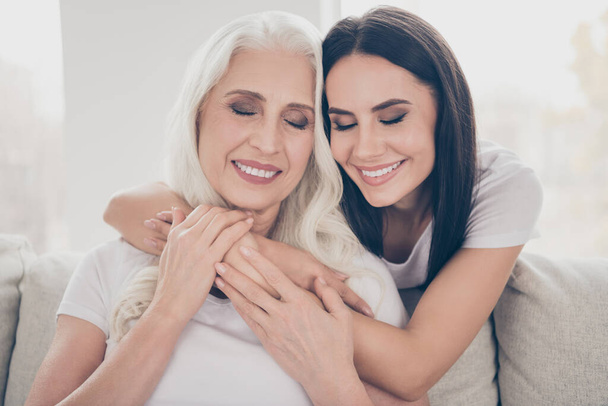 Close-up portrait of two nice attractive lovely cheerful dreamy sweet gentle women sitting on sofa granddaughter embracing grandma enjoying day time in white light interior house apartment - Photo, Image