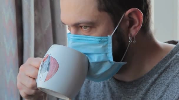 Coronavirus móka. Vicces férfi otthon, aki védőmaszkon keresztül teázik. Őrült otthon, férfi karanténban. Közelkép - Felvétel, videó