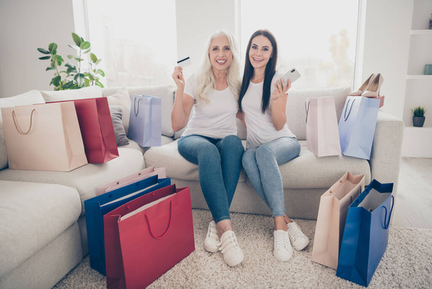 Portrait of two nice attractive lovely pretty cheerful cheery women ordering new clothes things trend web app delivery sitting on divan in light white interior room house flat apartment - Foto, afbeelding