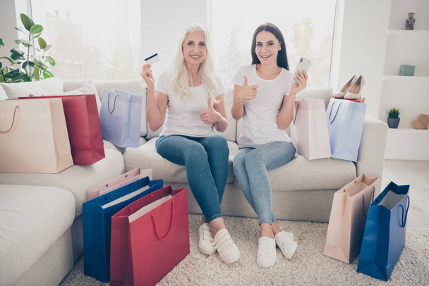 Portrait of two nice attractive lovely cheerful women shopper shopaholic sitting on sofa shopping online web app buying bags package purchase showing thumbup in white light interior house apartment - Foto, Imagen