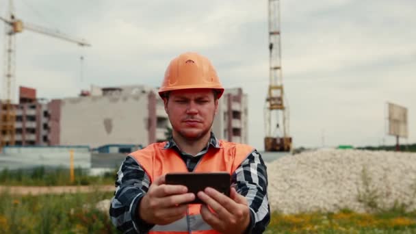 A man in an orange vest and orange helmet takes pictures of himself on a construction site on a mobile phone. - Кадры, видео