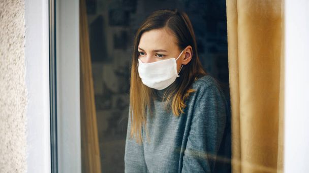 A woman in a gauze mask looks through the window. Anti-virus home isolation concept. Protect your health during the epidemic. - Fotografie, Obrázek
