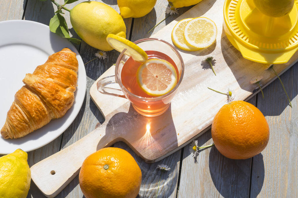 A cup of citrus fruit tea, croissant breakfast, lemons and oranges in the morning sunlight on wooden background - Photo, Image