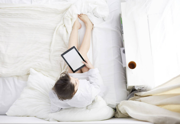 Portrait of woman using tablet and drinking coffee while sitting on the bed in the morning - Foto, afbeelding
