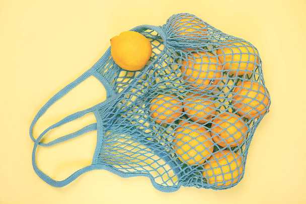 A lot of yellow lemon in the blue mesh bag flat lay on yellow background. Zero waste, eco friendly, no plastic concept. Citrus fruits. - Photo, image